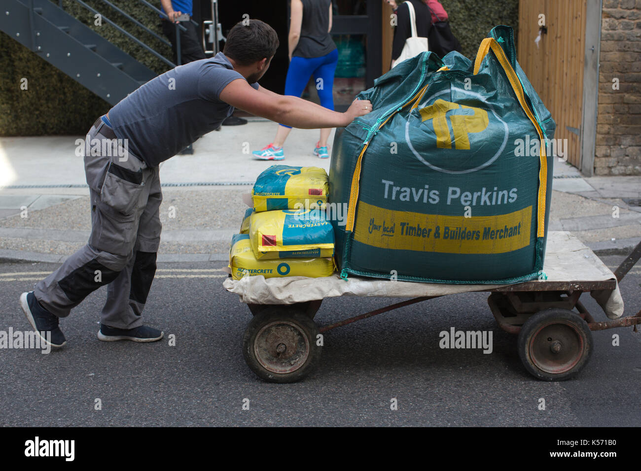 Uomo carrello di spinta con Travis perkins timber & builders merchant di cemento e sabbia per la costruzione, la zona centrale di Londra, Regno Unito Foto Stock