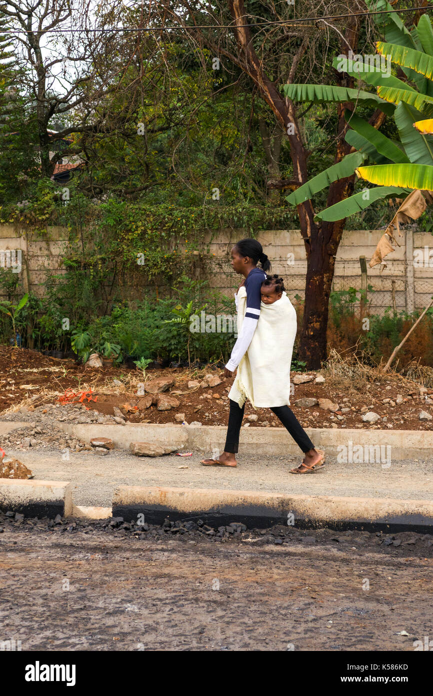 Donna bambino portando sul retro passeggiate sul marciapiede, Nairobi, Kenia Foto Stock