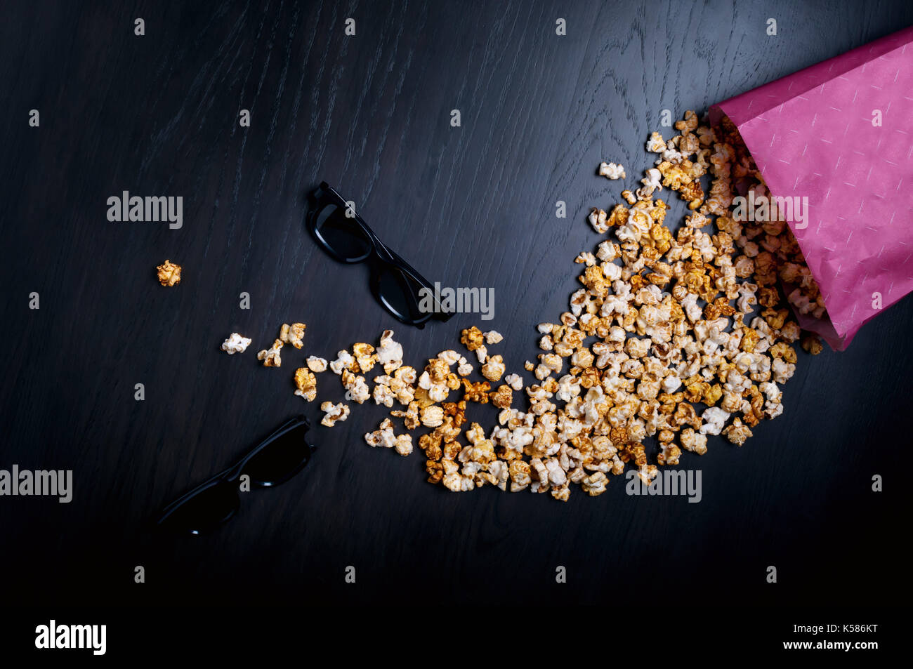 Popcorn sparsi sul legno scuro dello sfondo con paio di occhiali 3d. luminose movie argomento. fast food. rosa scatola di carta. la visione di cinema. Foto Stock