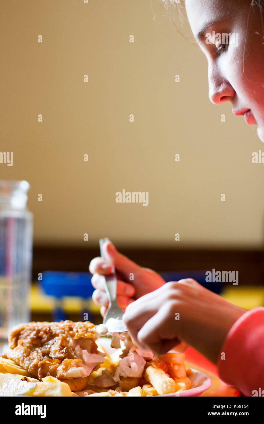 Ragazza di mangiare pesce e patatine con la lama di un coltello e forchetta, closeup, cena affamati, Foto Stock