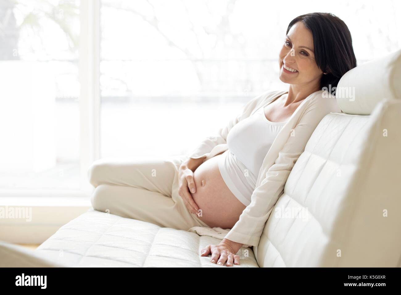Donna incinta seduta sul divano di toccare la pancia. Foto Stock