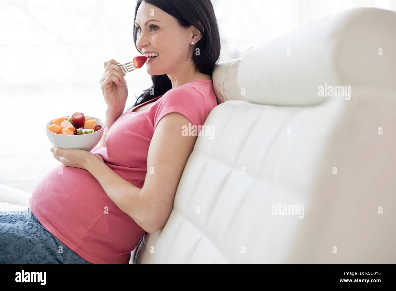 Donna incinta sul divano mangiando frutta fresca. Foto Stock