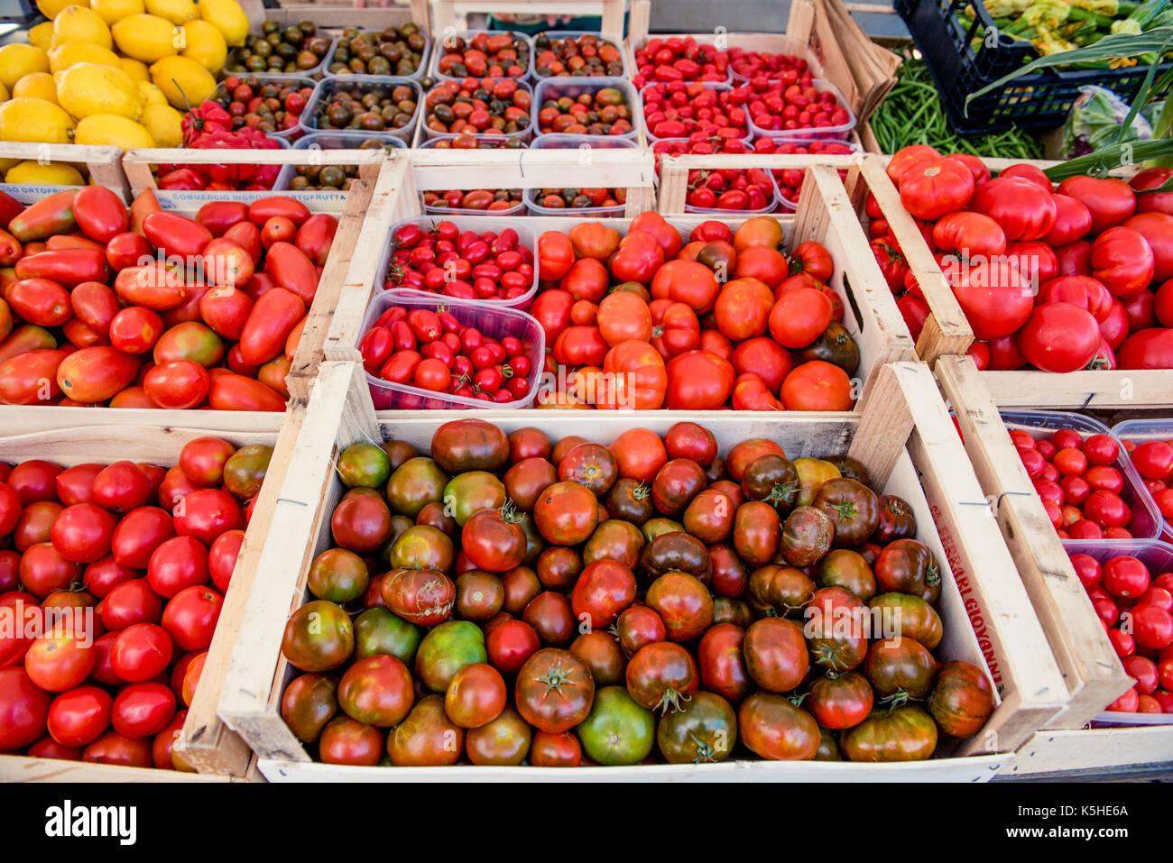 Pressione di stallo di mercato ad alto angolo di visione, casse di legno pieno di diverse varietà di pomodoro, limone e vegetali verdi in background Foto Stock
