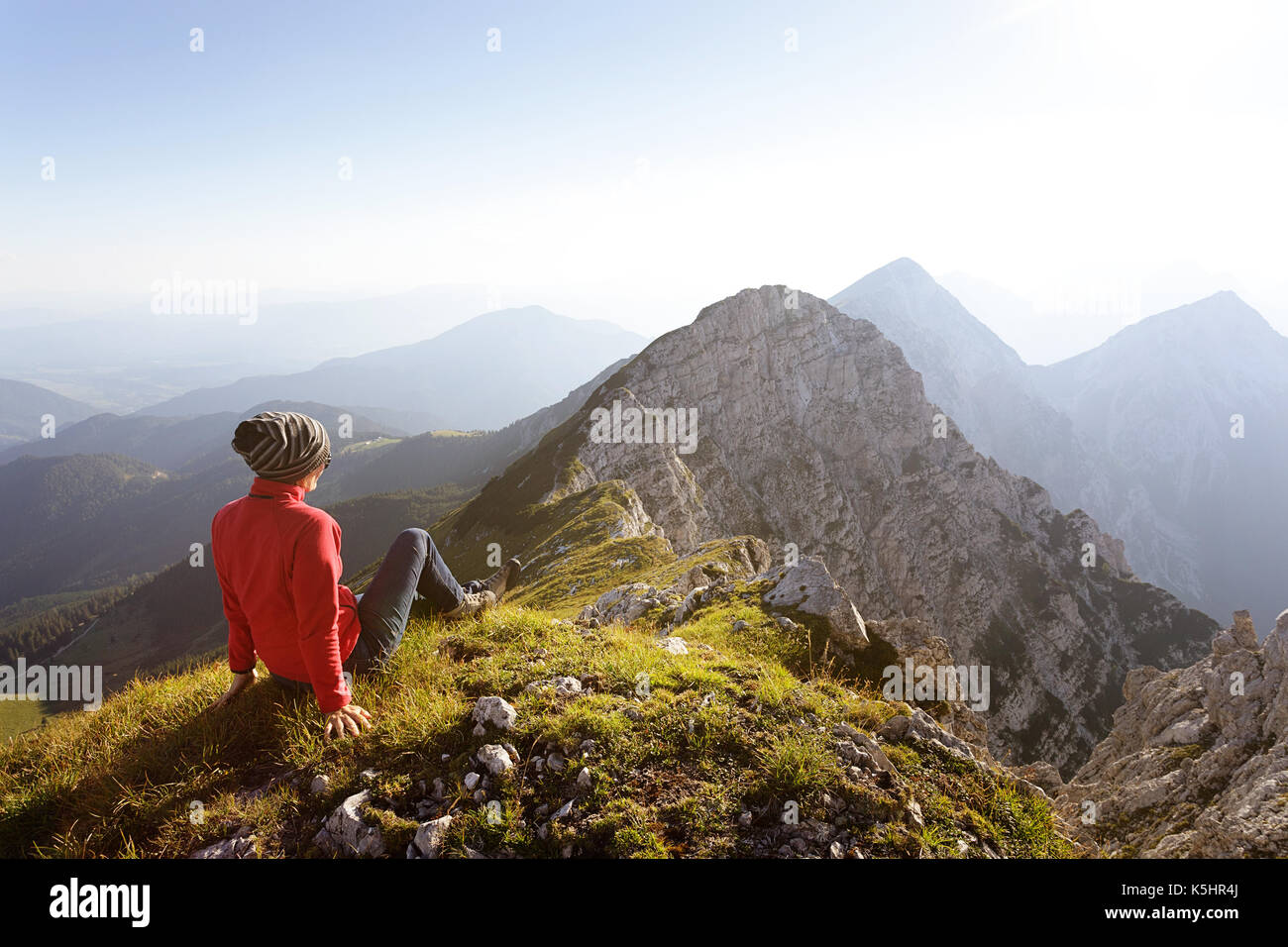 Woman in Red escursionismo jacket seduto sull'erba in cima alla montagna con vista mozzafiato sulle cime. Kladivo, Karakvanke, Slovenia. Foto Stock