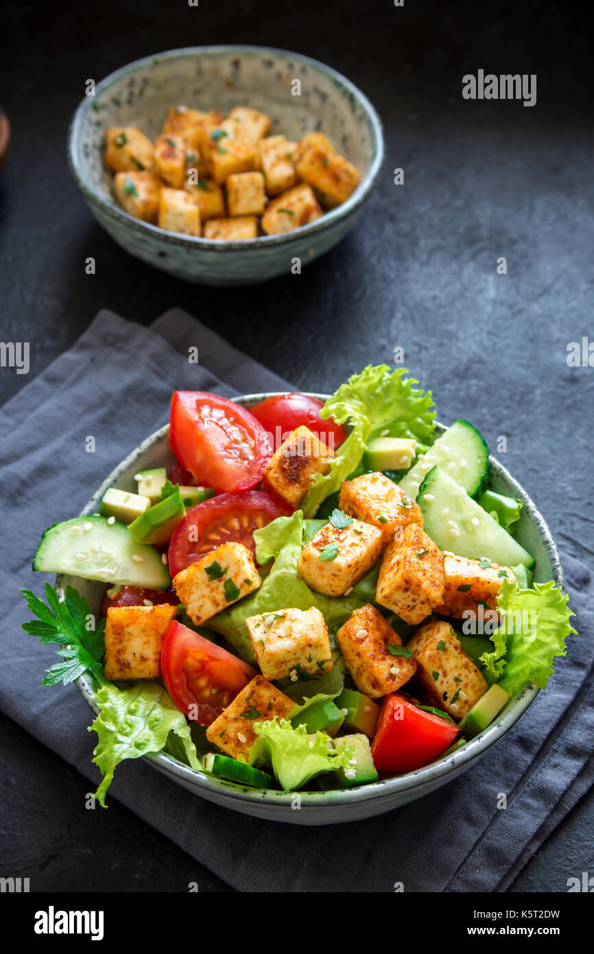Tofu fritto con insalata di cetrioli e pomodori, avocado e semi di sesamo. in casa asiatica di verdure e insalata di tofu in vaso in ceramica nera su sfondo di pietra Foto Stock