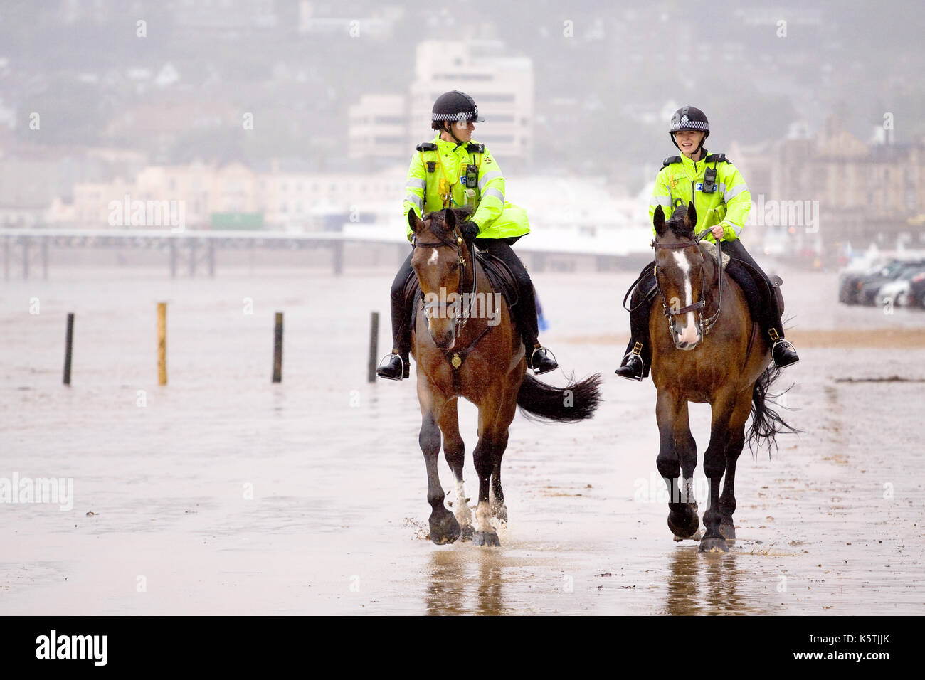 Due mouned donna di polizia, di pattuglia, in divise fluorescenti a cavallo.la loro polizia cavalli lungo una spiaggia in wet ventoso. Foto Stock
