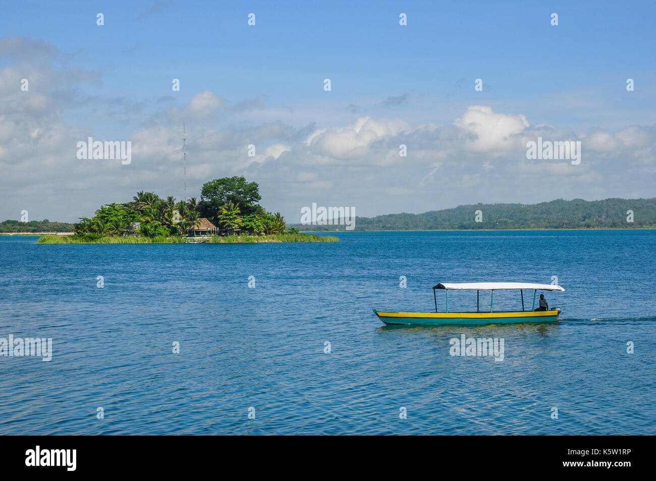Una piccola barca coperta galleggia su un calmo ed esteso specchio d'acqua con una lussureggiante isola verde sullo sfondo sotto un cielo parzialmente nuvoloso. Foto Stock