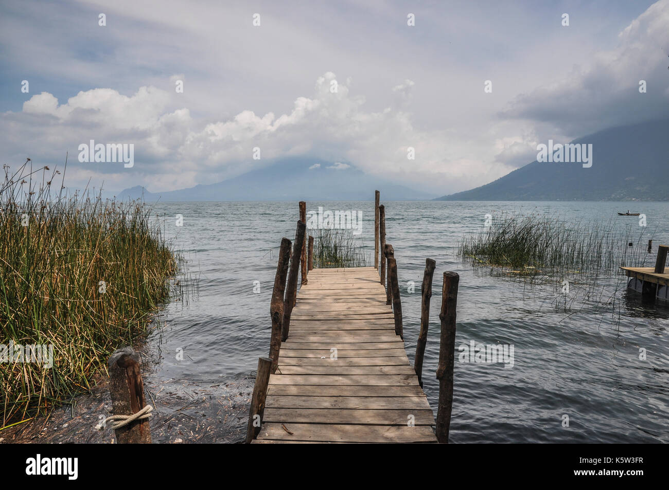 Un molo di legno si estende in un lago calmo circondato da montagne e sotto un cielo nuvoloso. Foto Stock