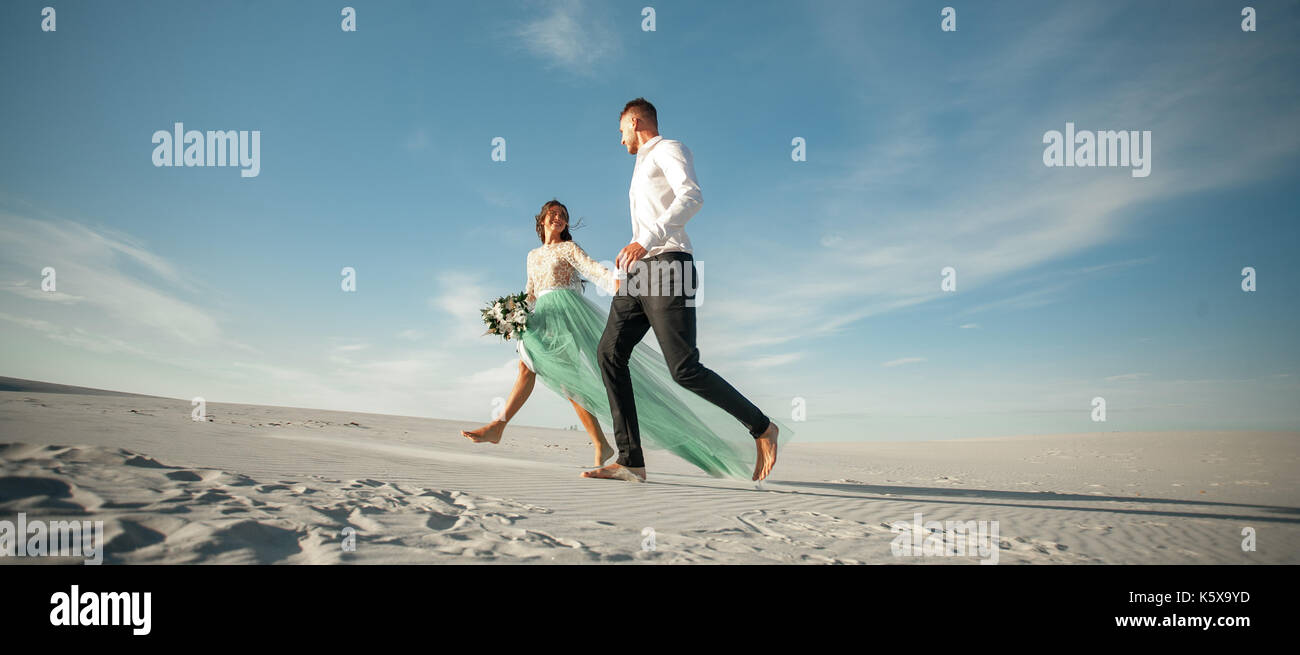 Sposa e lo sposo tenere mani, sorridere e camminare a piedi nudi nel deserto. sposa è vestito in abiti da sposa e con bouquet. Essi andando su sfondo bianco di s Foto Stock