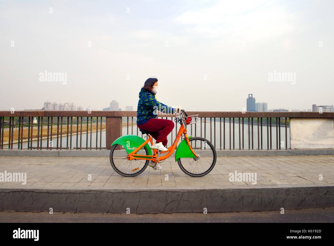 Città di Taiyuan, shanxi, Cina. giovane donna indossa l'inquinamento atmosferico maschera rides city community bike ciclo di quota sopra il fiume fen Foto Stock