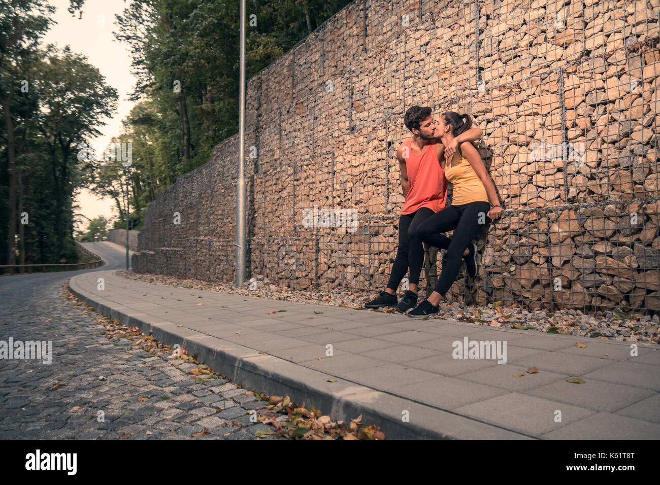Due giovani persone adulte, seduta rocce, all'aperto natura foresta asfalto indumenti sportivi, abbraccio bacio Foto Stock