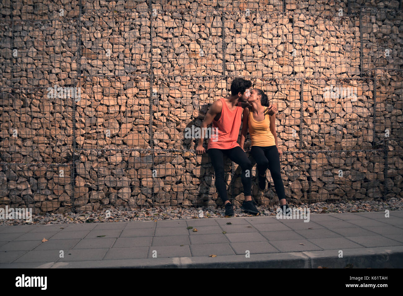 Due giovani persone adulte, seduta rocce, all'aperto natura foresta asfalto indumenti sportivi, abbraccio bacio, marciapiede Foto Stock
