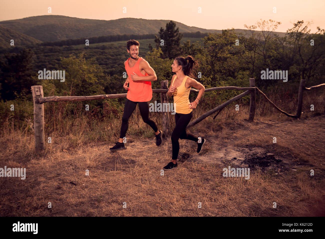 Due giovani, uomo donna, sorridenti, corridori correndo all'aperto, colori caldi, tramonto, montagne dietro Foto Stock