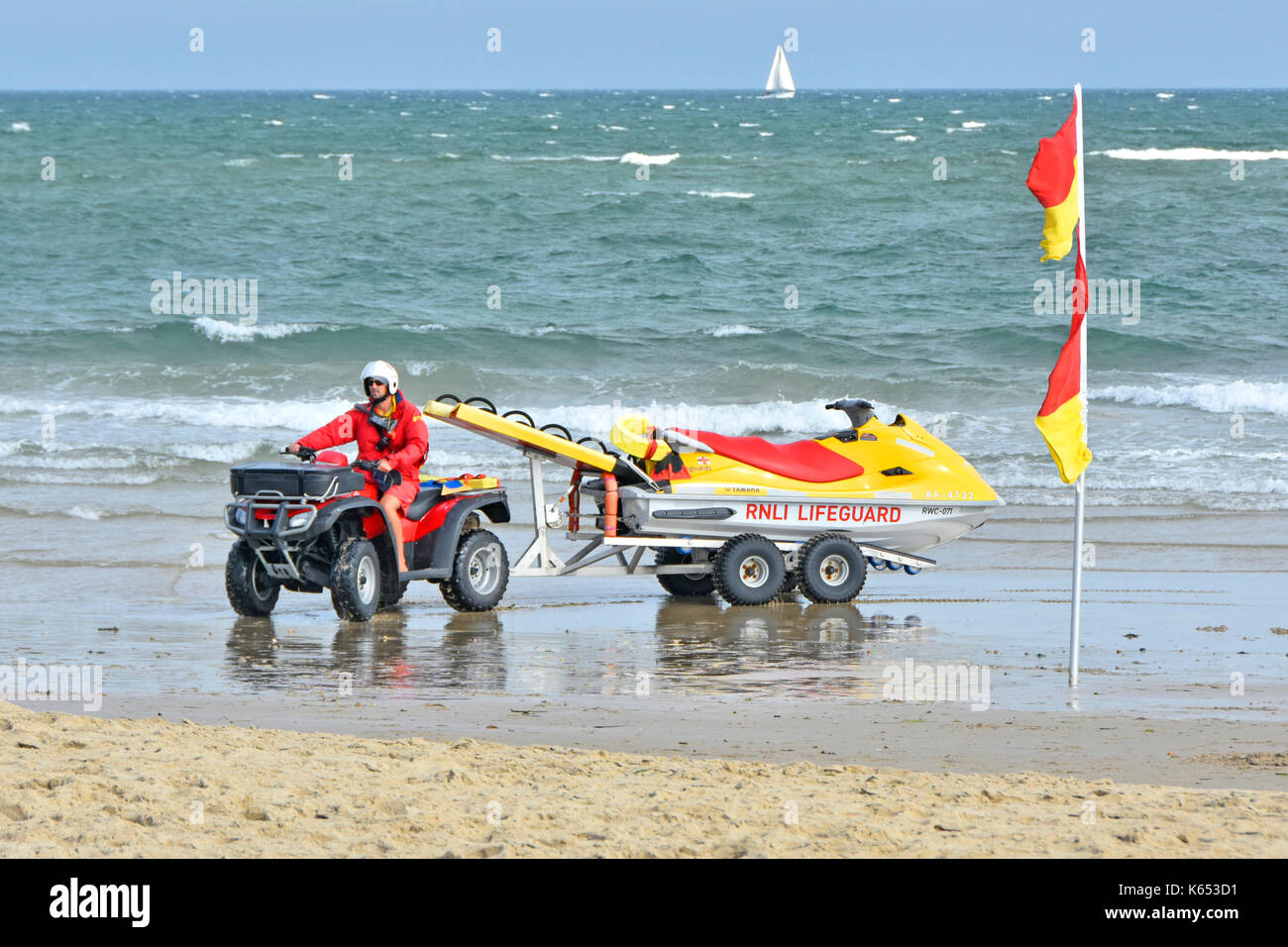 RNLI bagnino sul litorale a banchi di sabbia spiaggia alla guida di un ATV quad bike e Yamaha jet ski PWC mezzo di salvataggio con mare mosso e barca a vela distante Foto Stock