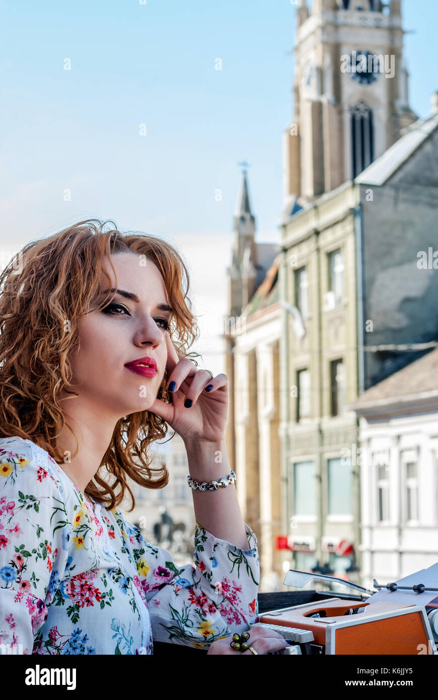 Giovane donna,look rétro, seduti a una macchina da scrivere, ritratto. rumore aggiunto. Una donna siede su di una terrazza al di sopra della città e guardando attentamente nel dis Foto Stock