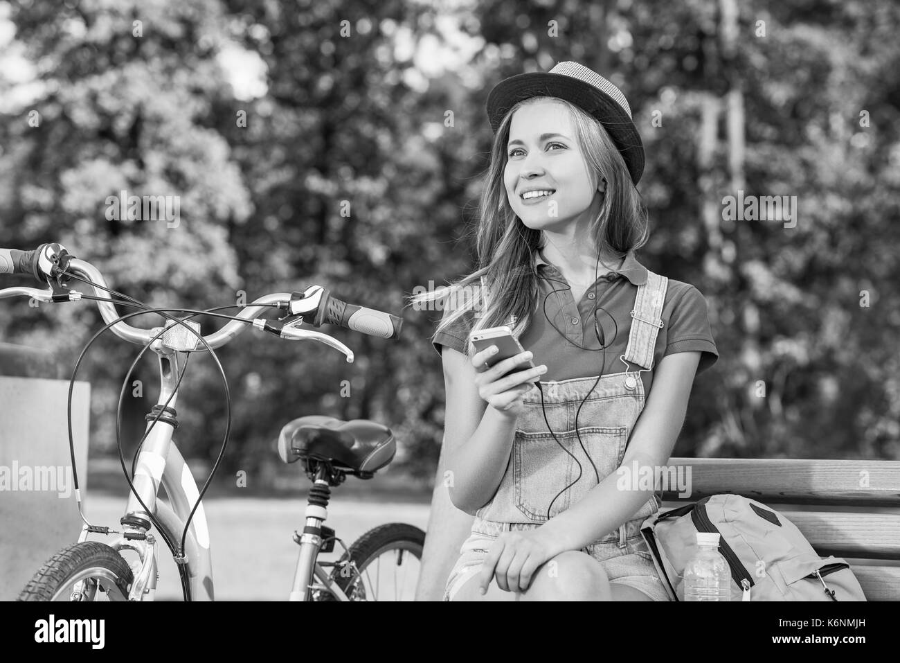 Girato in bianco e nero di una giovane e bella donna che indossa gli auricolari per ascoltare la musica sul suo smart phone seduto presso il parco locale dopo escursioni in bicicletta copyspace r Foto Stock