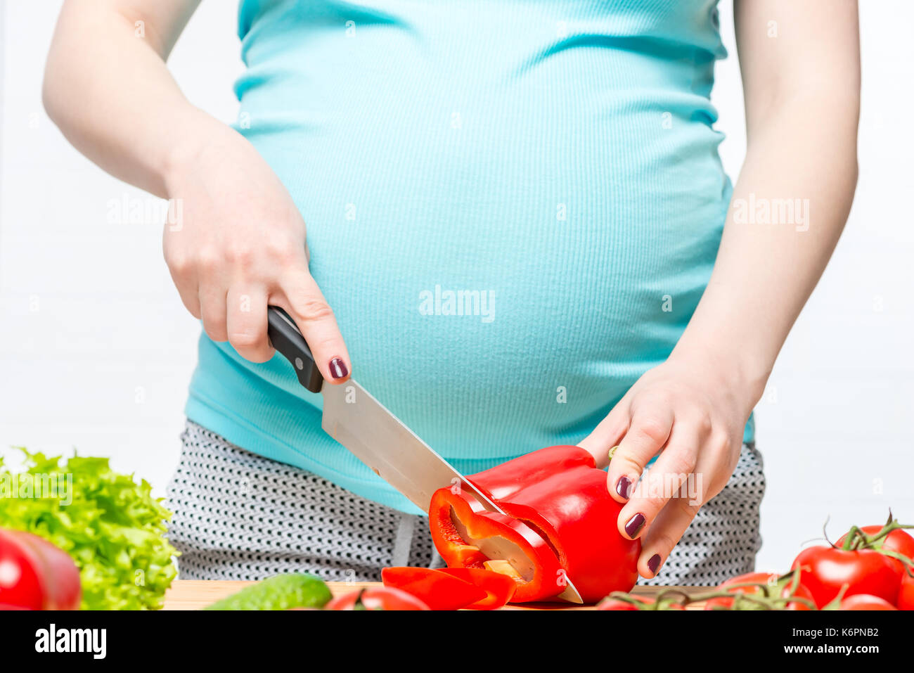 La pancia di una donna incinta di close-up e le mani con la lama di un coltello e verdure in cucina Foto Stock