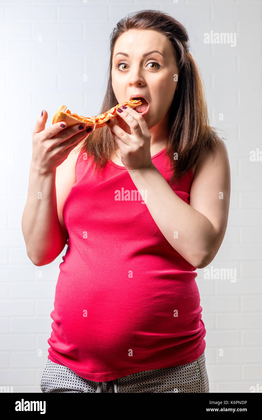 Fame donna incinta con una grande fetta di pizza su un mattone muro bianco sullo sfondo Foto Stock