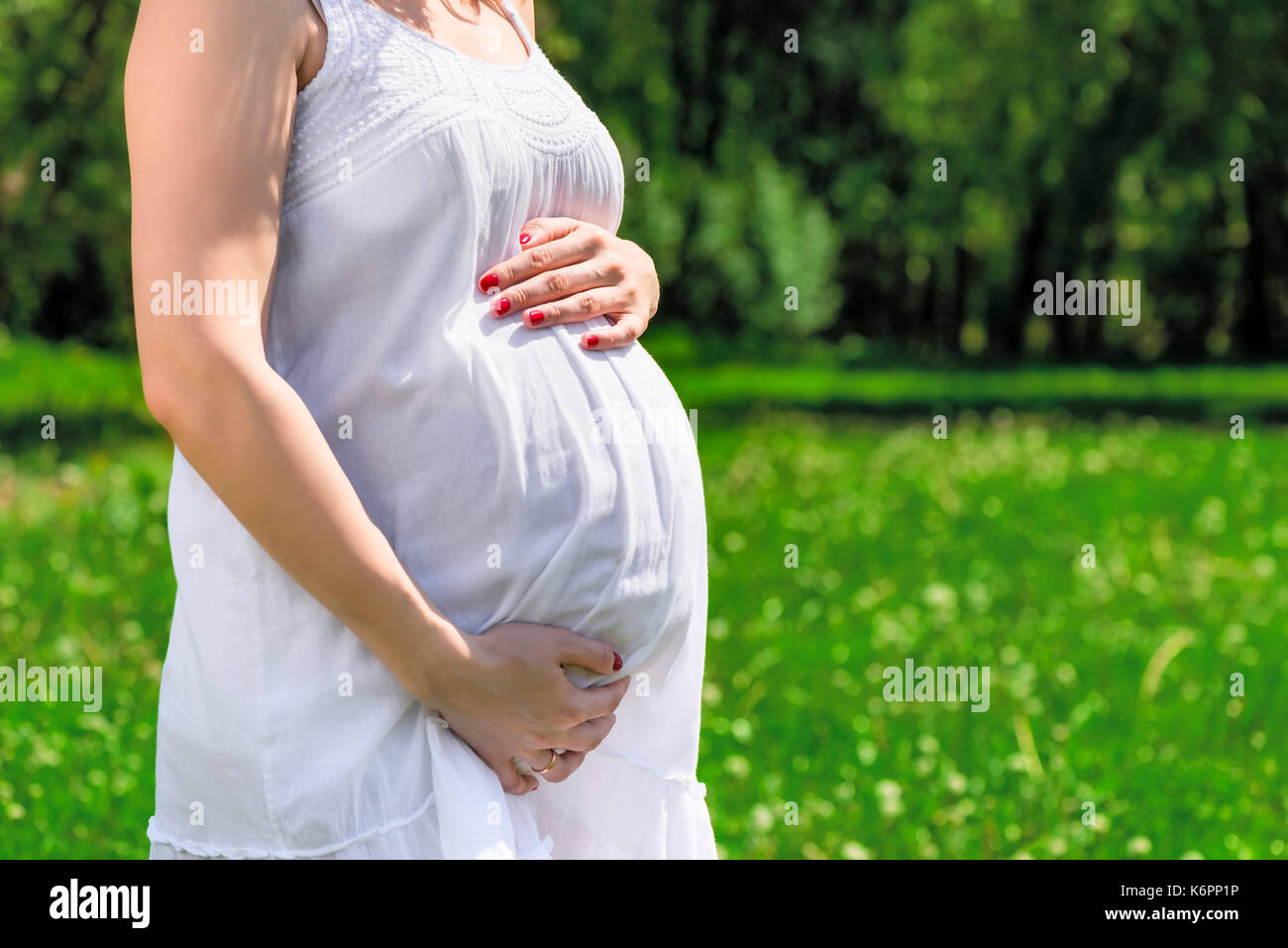 La pancia di una donna incinta in un abito bianco su uno sfondo di un parco verde Foto Stock