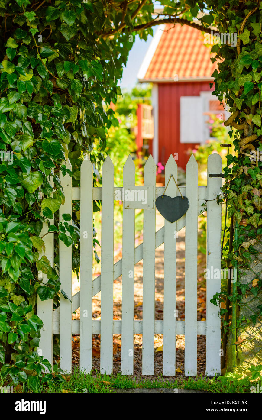 Legno bianco garden gate con cuore e numero di targa (numero rimosso). rosso cabina in legno sfocati sullo sfondo, hedgerows accanto al gate. Foto Stock