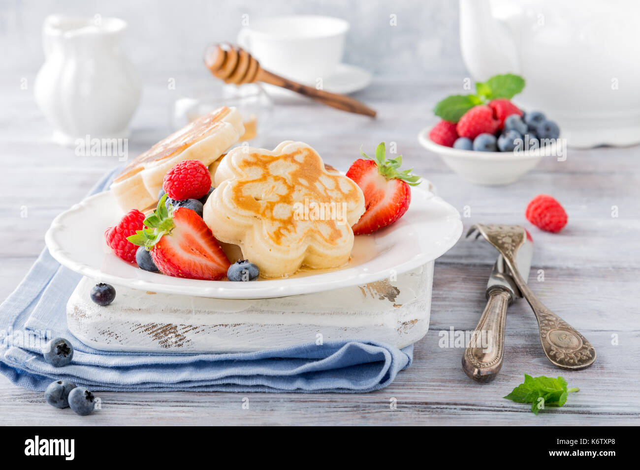 Frittelle di scotch in forma di fiori con frutti di bosco e miele bianco su sfondo di legno. colazione salutare concetto. Foto Stock