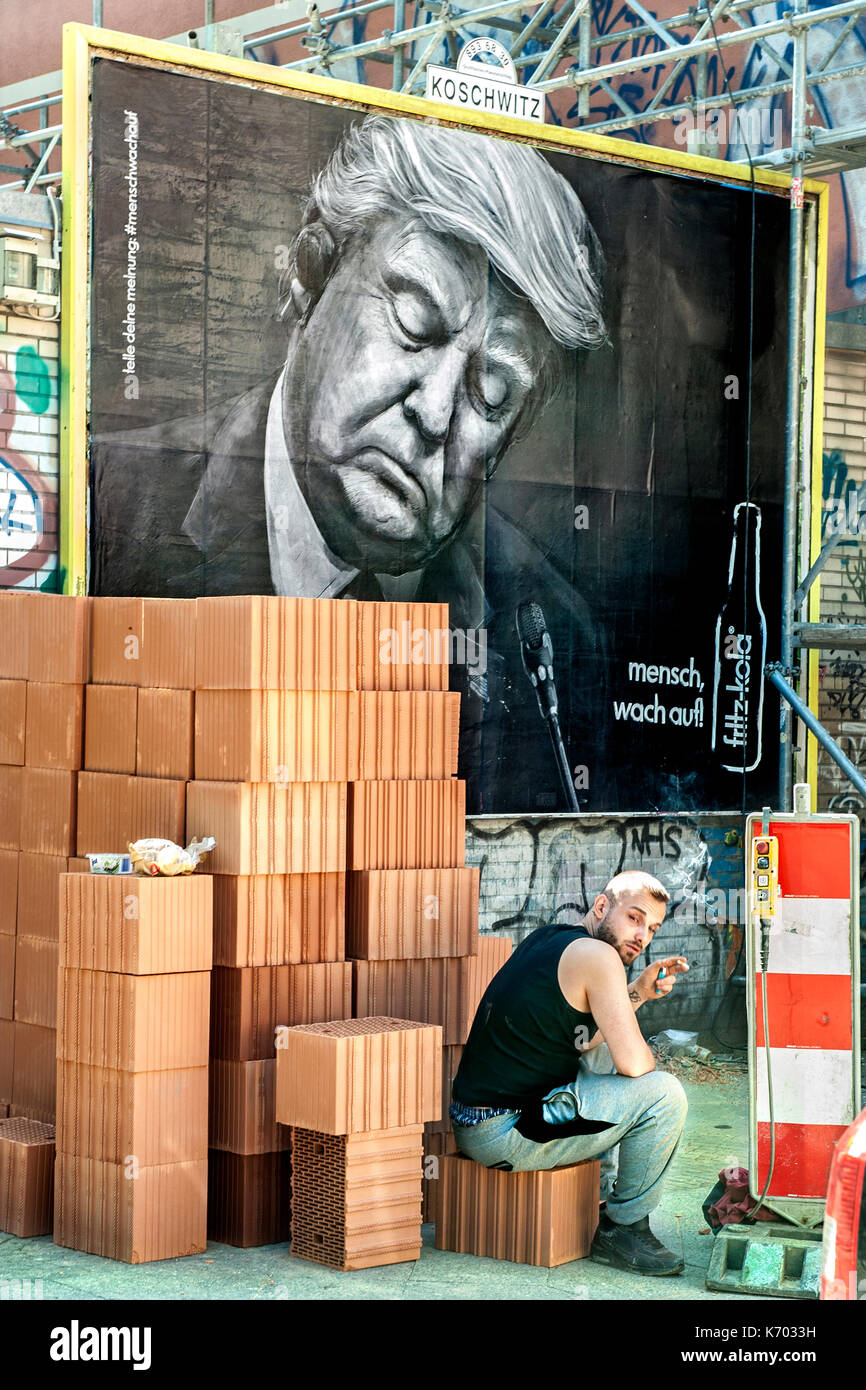 Germania UN muratore fuma mentre sta sollevando un muro di fronte a un poster pubblicitario con la foto del presidente Trump, a Schlesisches Tor, Berlino Foto Stock