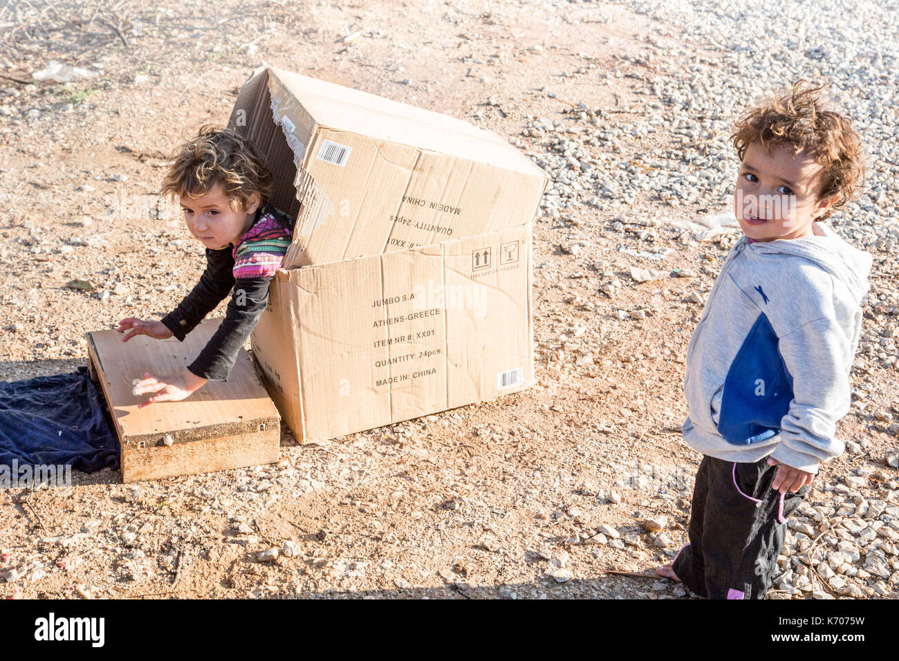 Un bambino gioca felicemente in una scatola di cartone sul fosco pietraie di ritsona Refugee Camp in Grecia come il suo amico guarda la telecamera. Foto Stock
