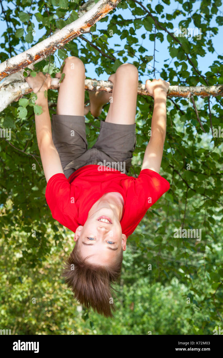 Basso angolo vista di felice ragazzo adolescente indossando t-shirt rossa appeso a testa in giù da un albero di betulla guardando la telecamera sorridendo godendo di estate Foto Stock