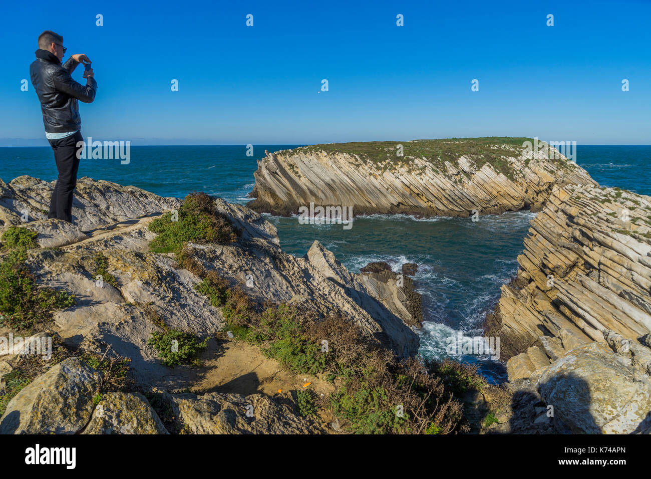 Uomo in piedi in cima a una scogliera di scattare una foto delle onde e la riva al di sotto del Portogallo Foto Stock