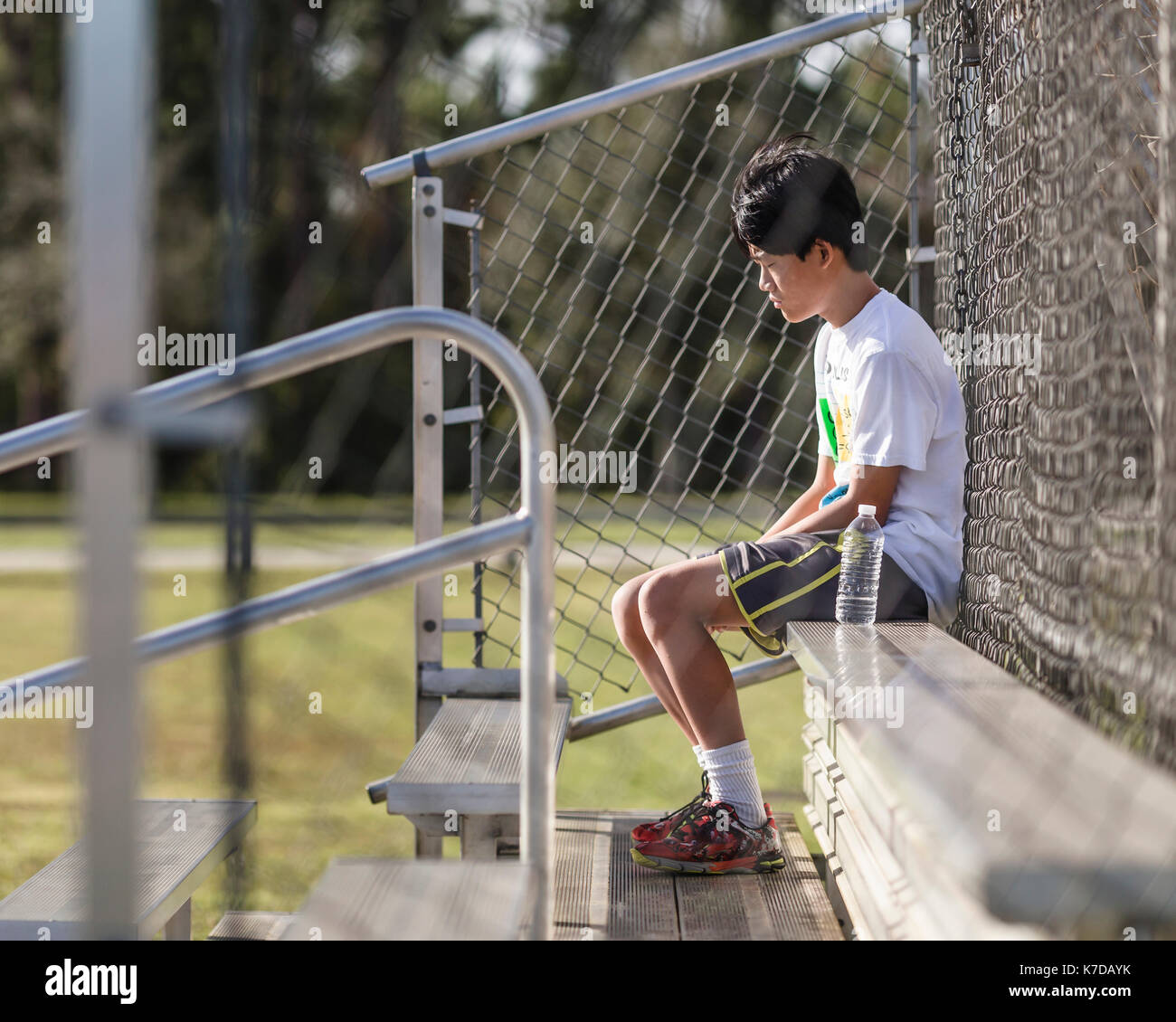 Riflessivo ragazzo adolescente seduto sulla panchina contro opaco recinzione al campo da gioco Foto Stock
