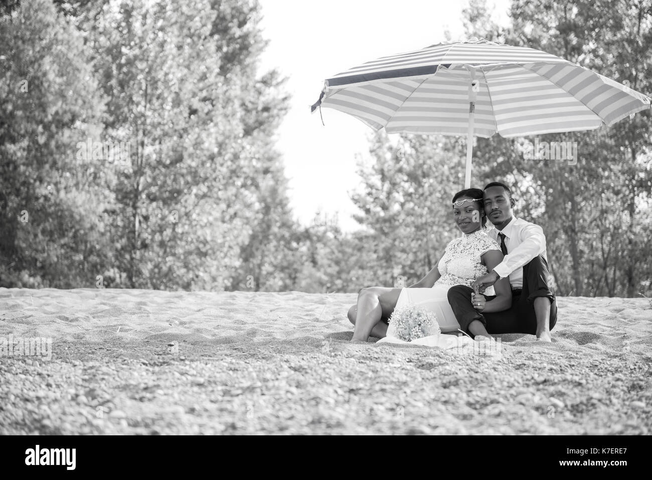 Giovane sposa e lo sposo seduta nella sabbia in spiaggia sotto ombrellone Foto Stock