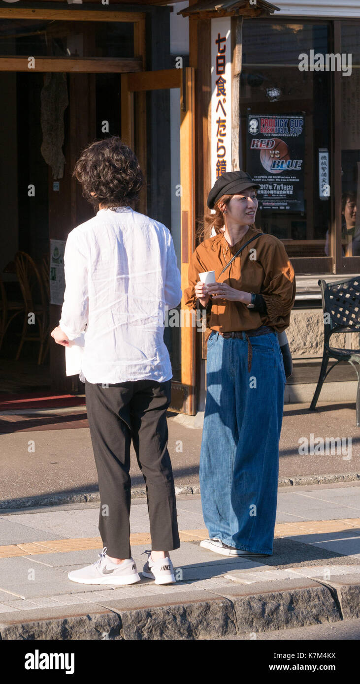 Un giovane giapponese giovane vestito in denim alla moda , bianco e nero camicia nel tardo pomeriggio di sole in piedi sul marciapiede che guarda lontano da e a telecamera. Foto Stock