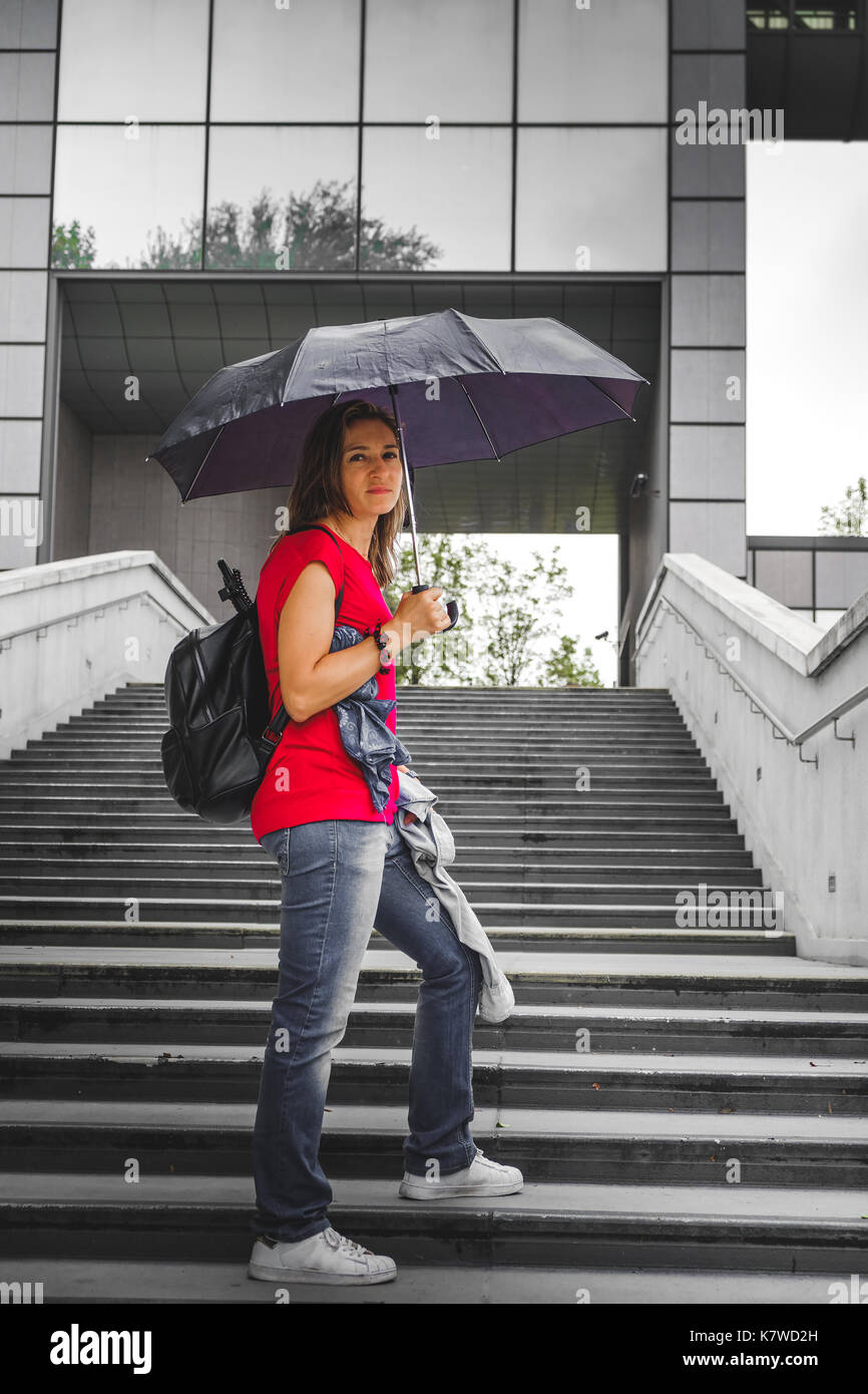 Tourist donna triste infelice a causa della pioggia in una grande città mentre salgono i gradini di trattenimento di un ombrello in un moderno quartiere Foto Stock