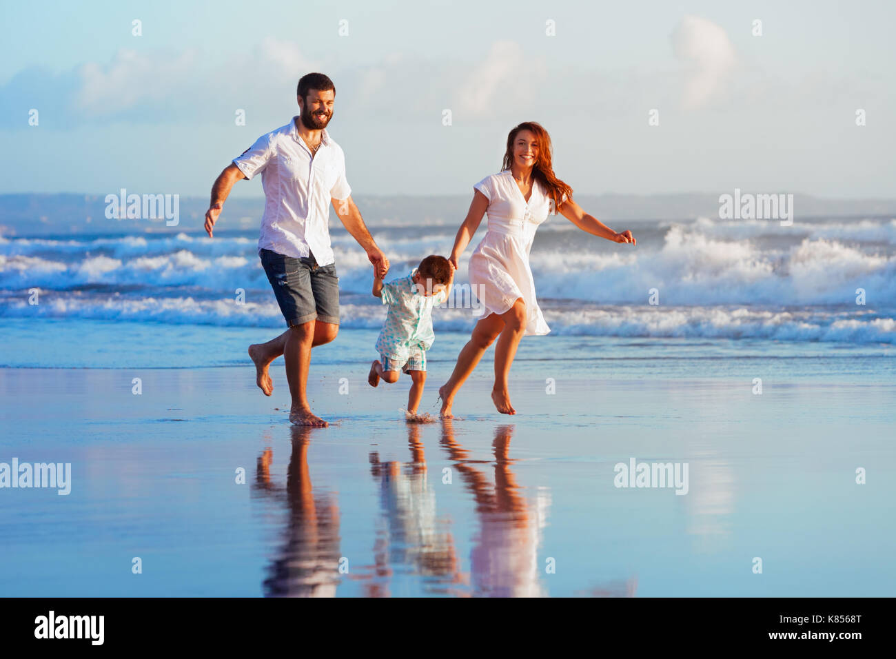 La famiglia felice - padre, madre, figlio bambino divertirsi insieme, bambino correre con spruzzi di acqua piscina lungo sunset sea surf sulla spiaggia di sabbia nera. Foto Stock