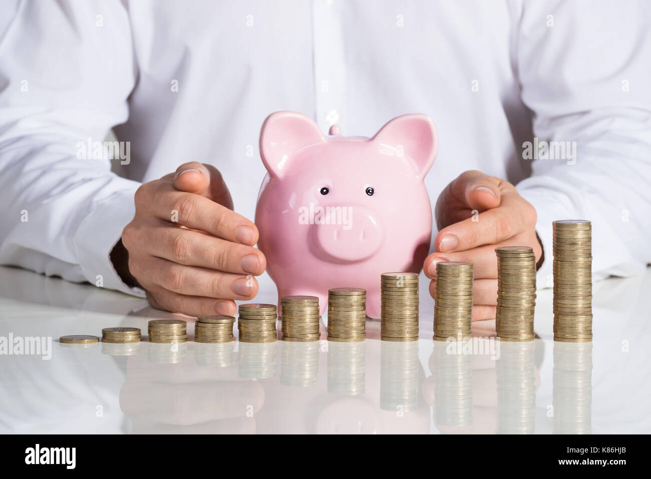 Sezione mediana di imprenditore proteggere piggybank con monete impilate disposte a scrivania da ufficio Foto Stock
