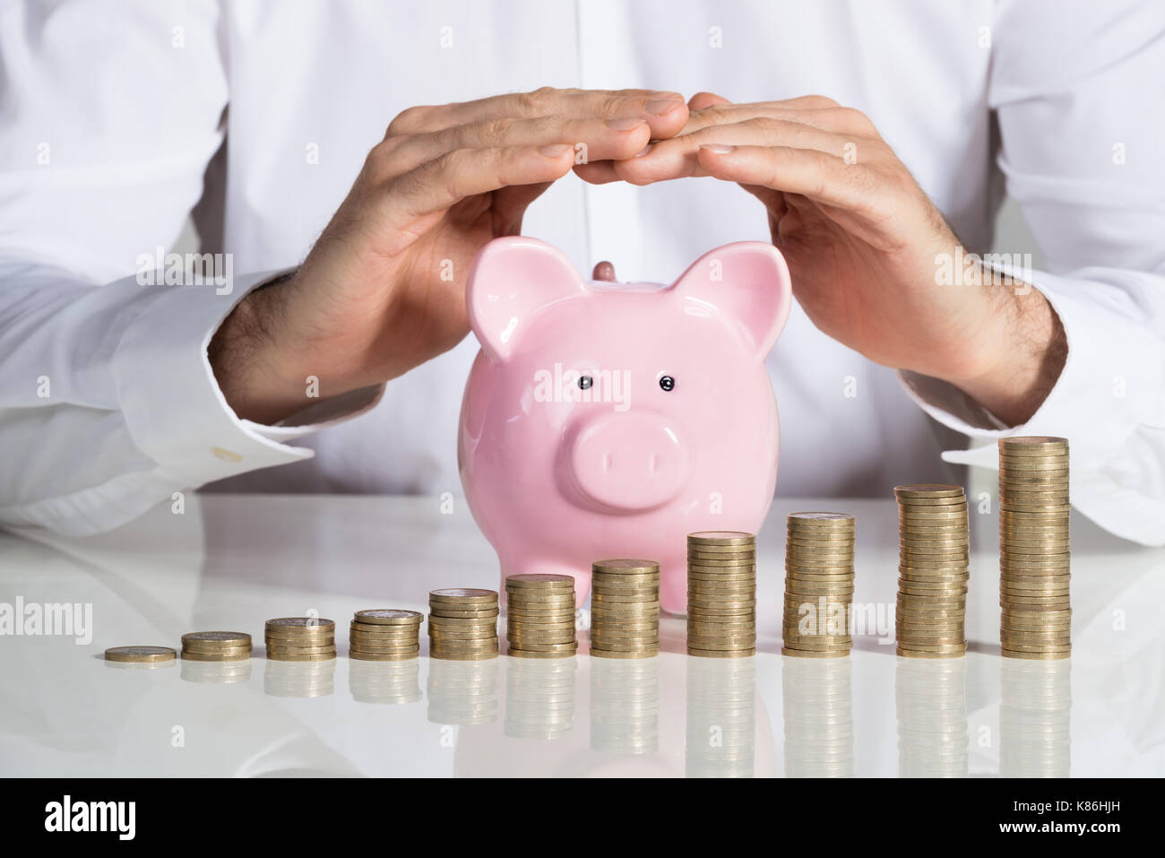 Sezione mediana di imprenditore proteggere piggybank con monete impilate disposte a scrivania da ufficio Foto Stock