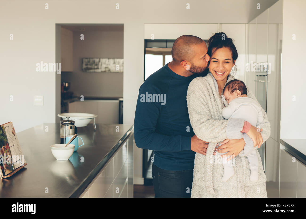 Uomo di baciare la moglie tenendo un neonato ragazzo in cucina. Bella giovane famiglia di tre di mattina in cucina. Foto Stock