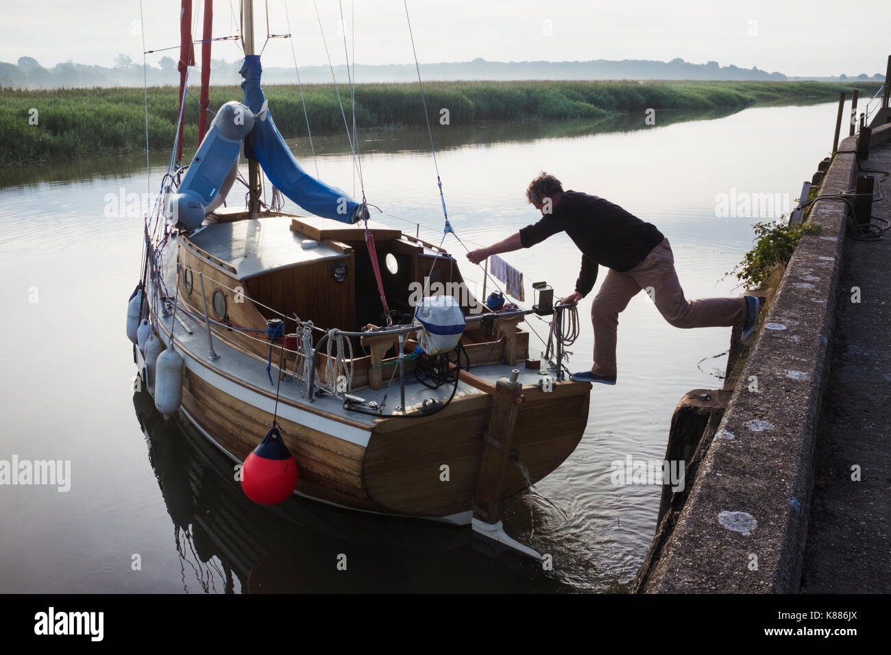 L'uomo bilanciamento tra la parete del porto con un piede su una barca a vela, spingendo la barca lontano dagli ormeggi nel canale d'acqua. Foto Stock