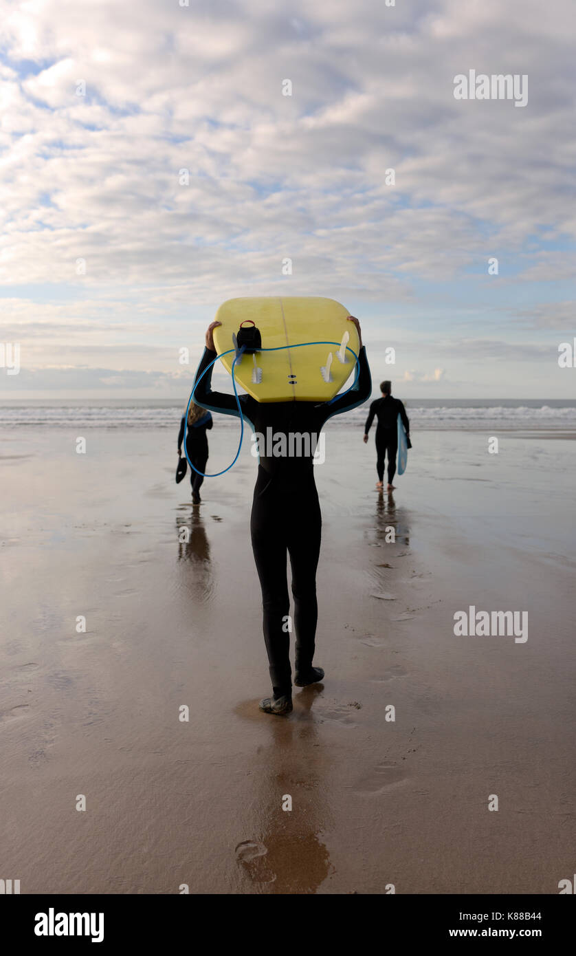 Una famiglia si prepara per il surf che trasportano le tavole da surf in tutta la spiaggia di indossare muta su una giornata d'estate. Foto Stock