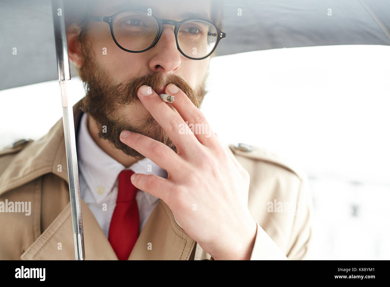 Elegante e chic uomo in cravatta rossa e beige trench di fumare sotto ombrellone Foto Stock
