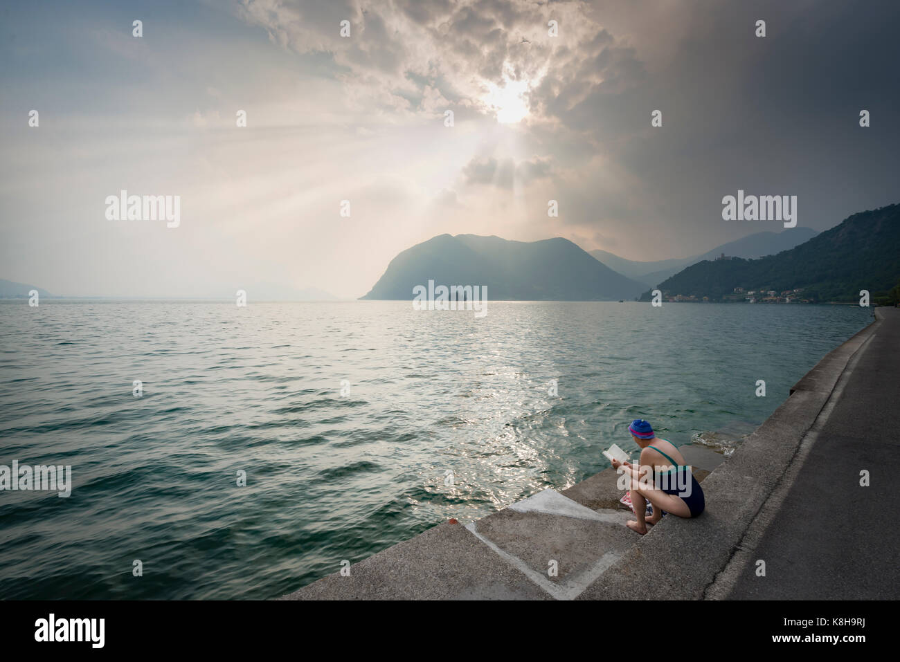 Donna seduta in un costume da bagno e la lettura sul banco di monte isola nel lago d'Iseo gli ultimi raggi di sole prima di un temporale in salita, Italia Foto Stock