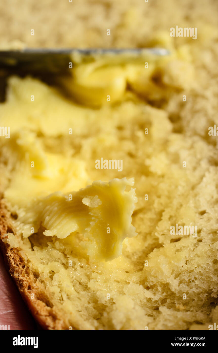 Una fetta di fresca, pane caldo essendo imburrato con coltello in soft focus sullo sfondo e la piccola parte di mano azienda è appena visibile. Foto Stock
