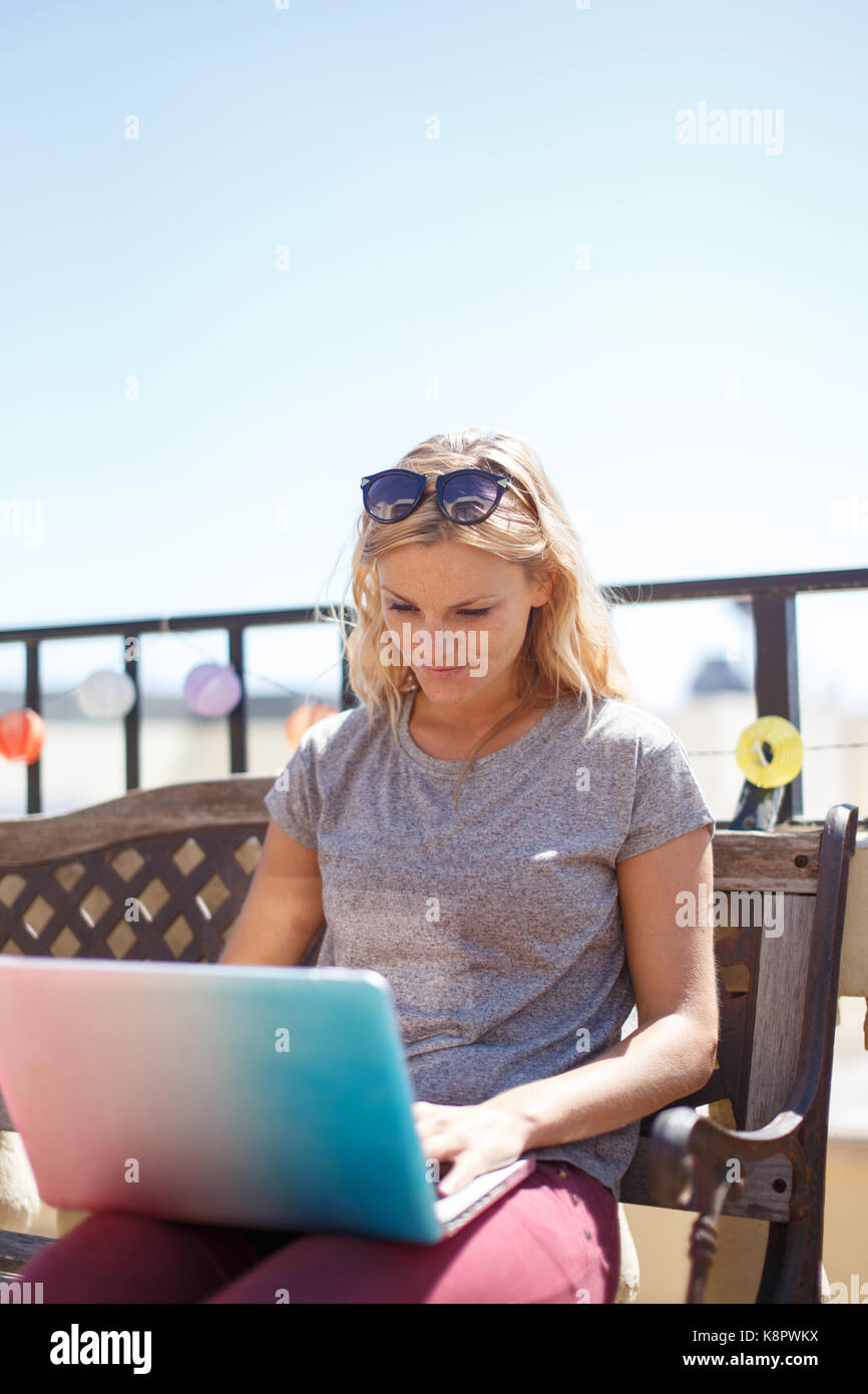 Giovane donna bionda digitando su laptop seduta sul banco di prova all'aperto Foto Stock