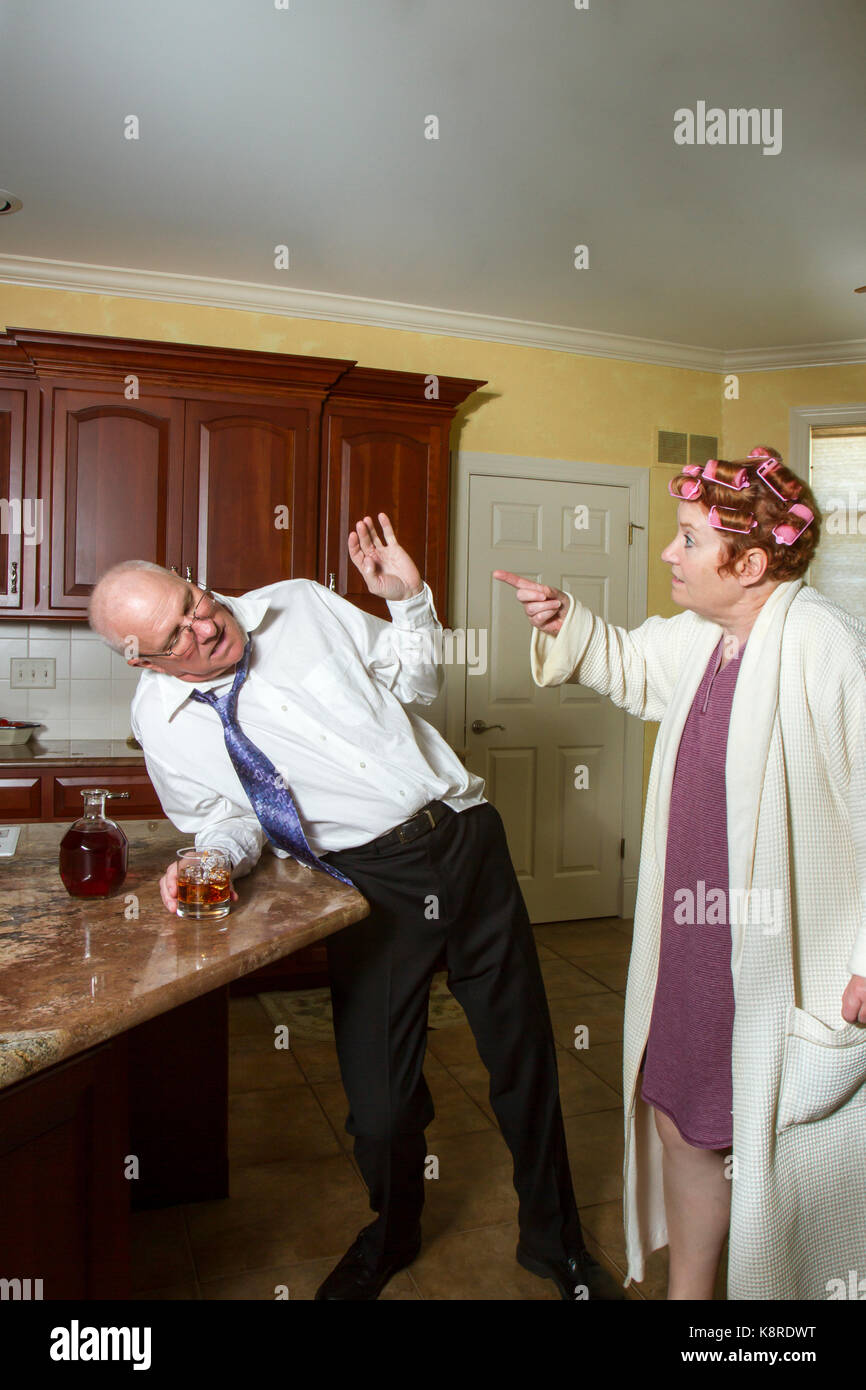 Uomo ubriaco appoggiato contro il contatore in cucina con arrabbiato moglie in accappatoio e arricciacapelli urlare. Foto Stock