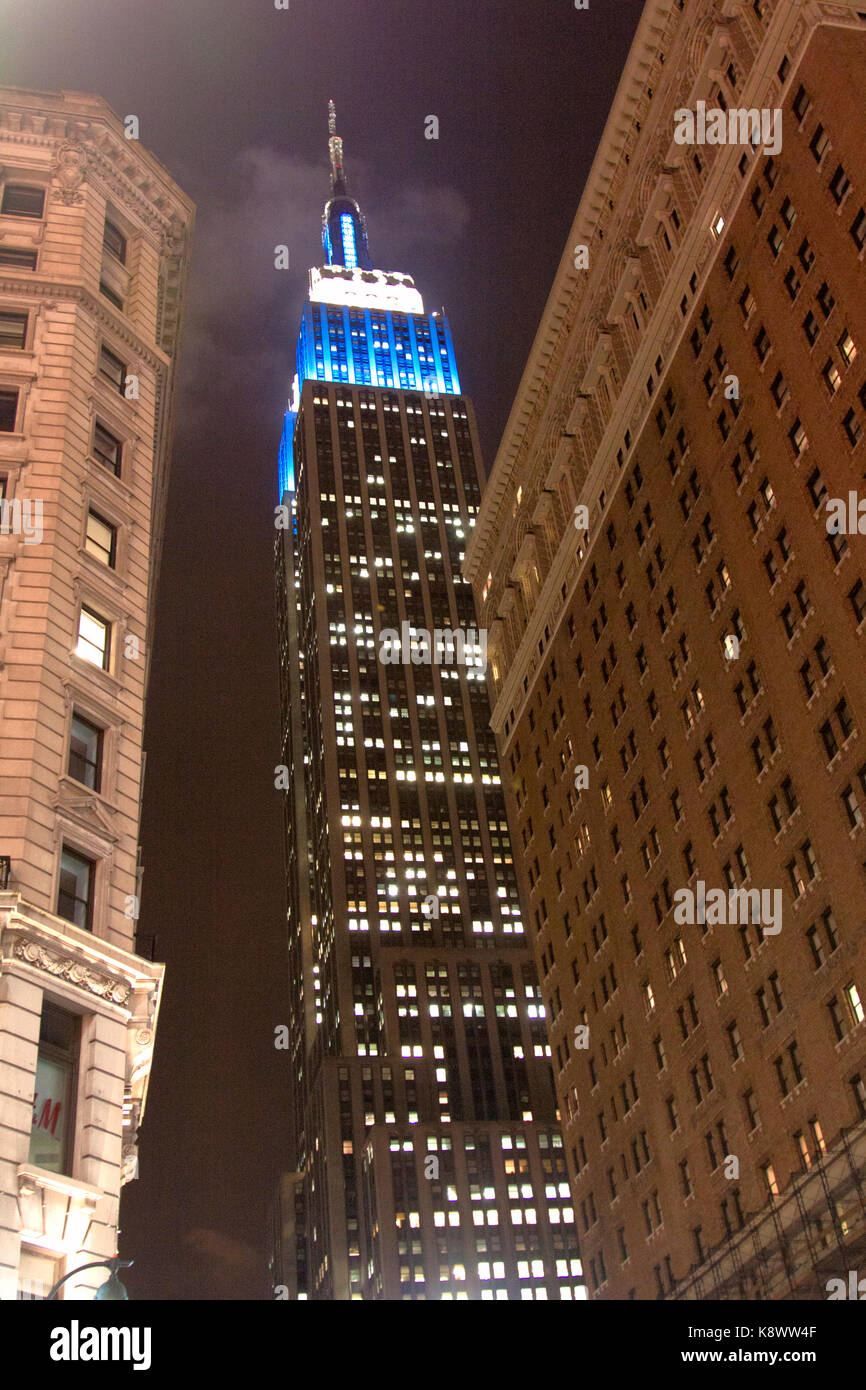 Una foto che guarda l'Empire state Building di notte dal livello della strada. La parte superiore è illuminata da luci blu e avvolta in una nuvola bassa. Foto Stock
