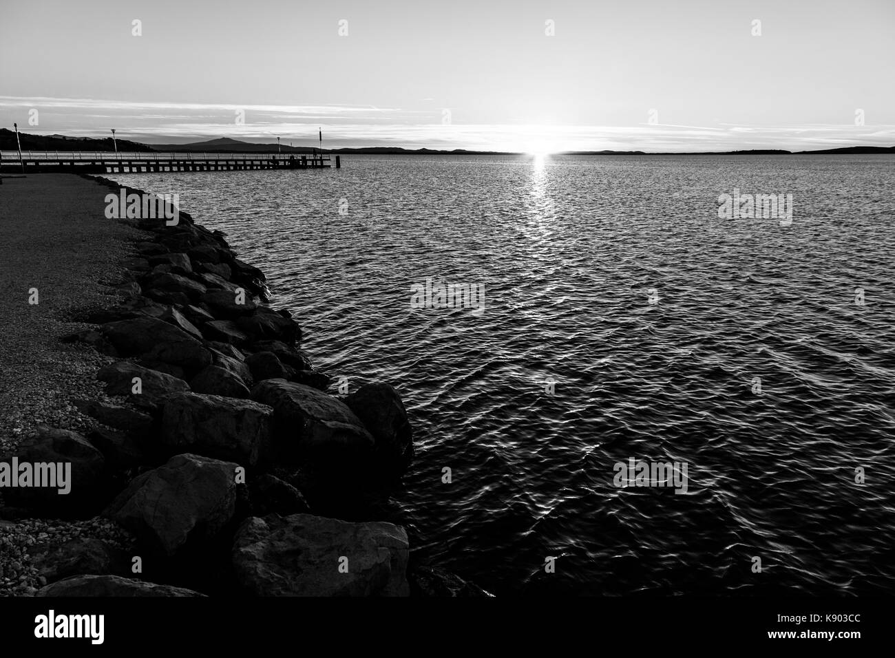 Tramonto sul lago, con il sole basso sull'orizzonte e rocce in primo piano Foto Stock