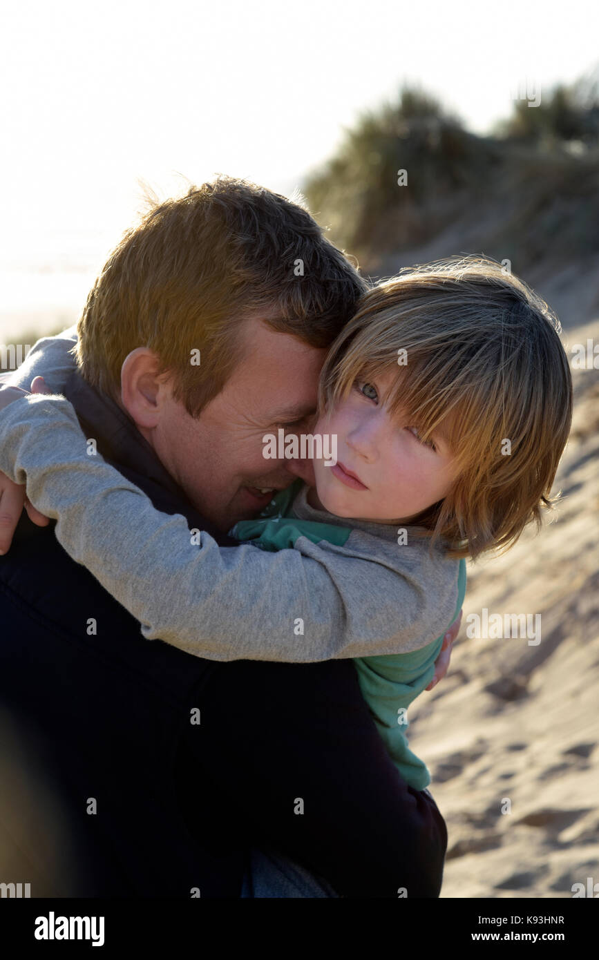 Un padre abbraccia il suo figlio sulle dune di sabbia in una giornata di sole, ragazzo guarda alla telecamera come suo padre coccole e abbracci lui vicino. Foto Stock