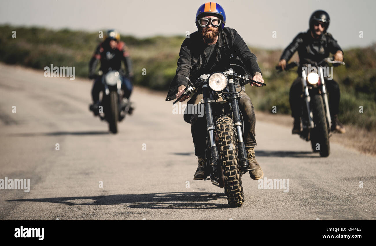 Tre uomini che indossano faccia aperta caschi e occhiali in sella cafe  racer moto lungo la strada rurale Foto stock - Alamy