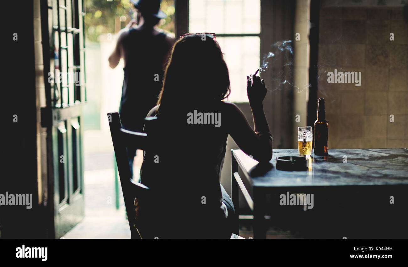 Vista posteriore silhouette di donna seduta al chiuso in una tabella, il fumo di sigarette, bicchiere di birra e una bottiglia sul tavolo. Foto Stock