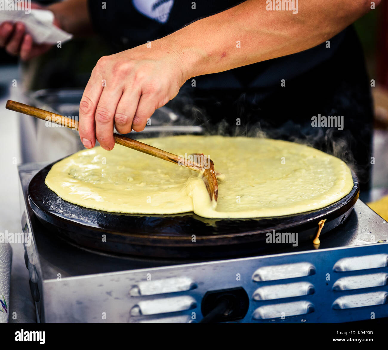 Realizzazione di Crepes Pancake nel mercato aperto festival fiera. una mano è rendere crepes all'aperto su una griglia metallica con bastone di legno su un outdoor summer fest Foto Stock